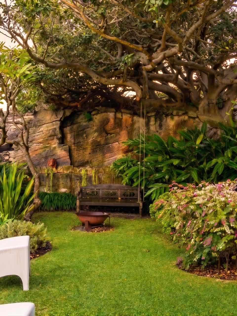 A sheltered Lüdi Gardens courtyard against a sandstone wall, with fig tree, bench swing and fire bowl