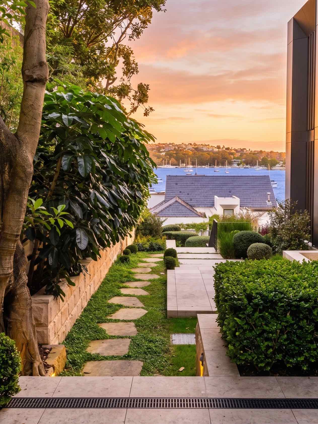 A terraced Lüdi Gardens landscape with a stone path and native planting framing a harbour view at sunset