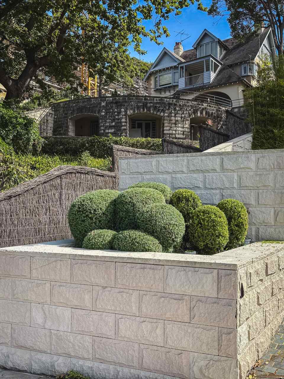 Cloud-pruned shrubs and clipped balls in a raised sandstone block planter below a terraced heritage garden