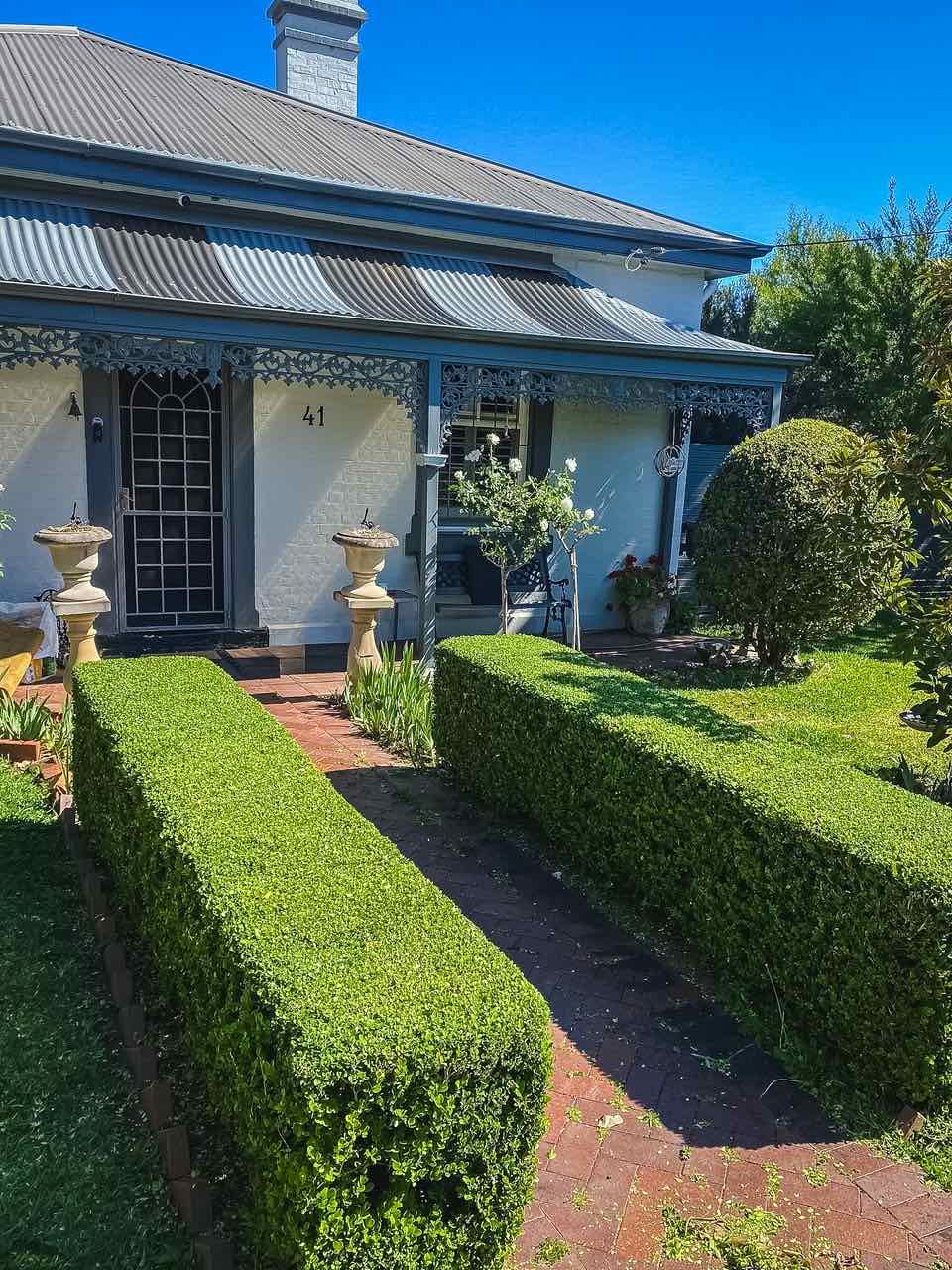 Freshly clipped box hedges along a brick path to a heritage cottage verandah with iron lacework