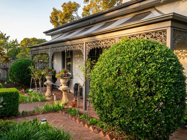 Rounded shrub and low hedges beside a zigzag-pattern brick path, in front of a white cottage veranda with decorative ironwork and stone pots of flowers